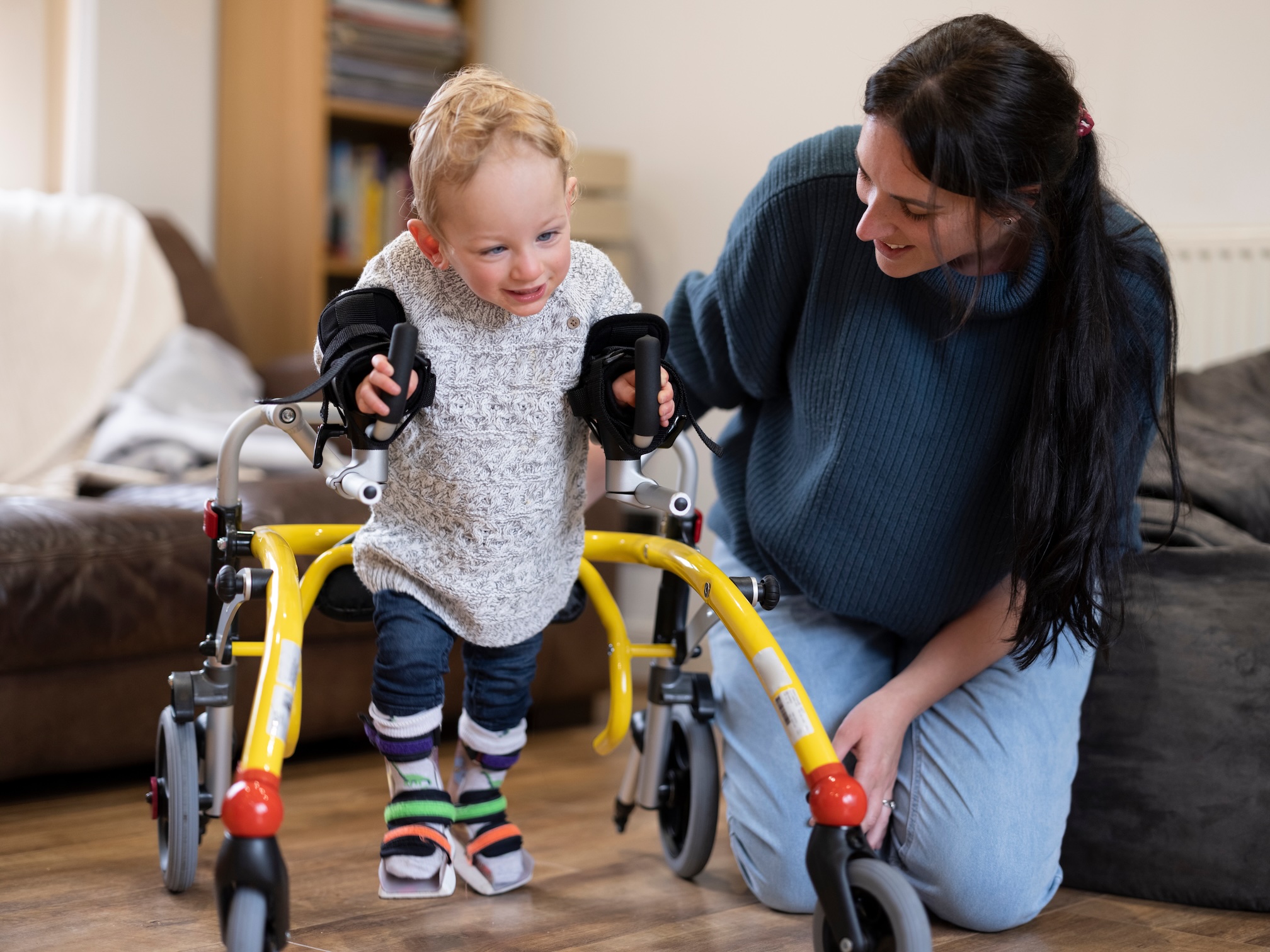 Mother encourages her toddler who has mobility impairments as he uses rolling walker to walk across floor at home.